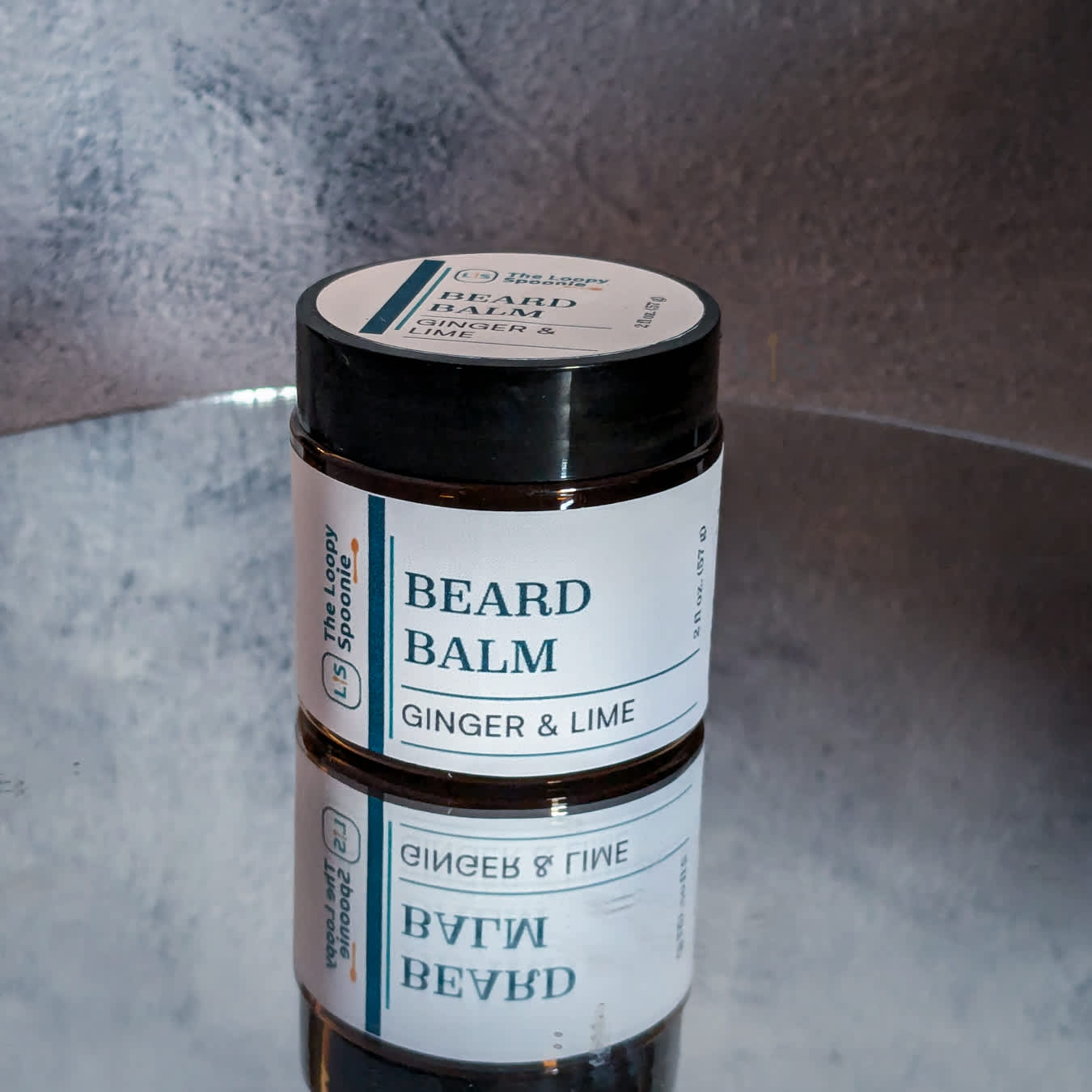 Dark amber jar sitting upright on a mirror against a grey background. The jar has a cream colored wrap around label that says Beard Balm, then below that "Ginger Lime."