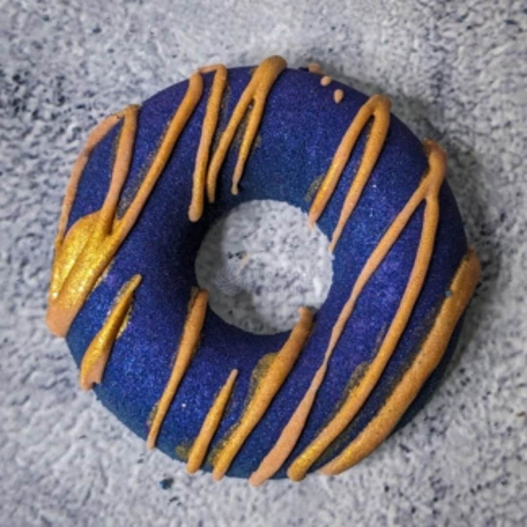 Royal blue donut bath bomb with gold/yellow cocoa butter drizzled on top.
The bath bomb is on a grey concrete background