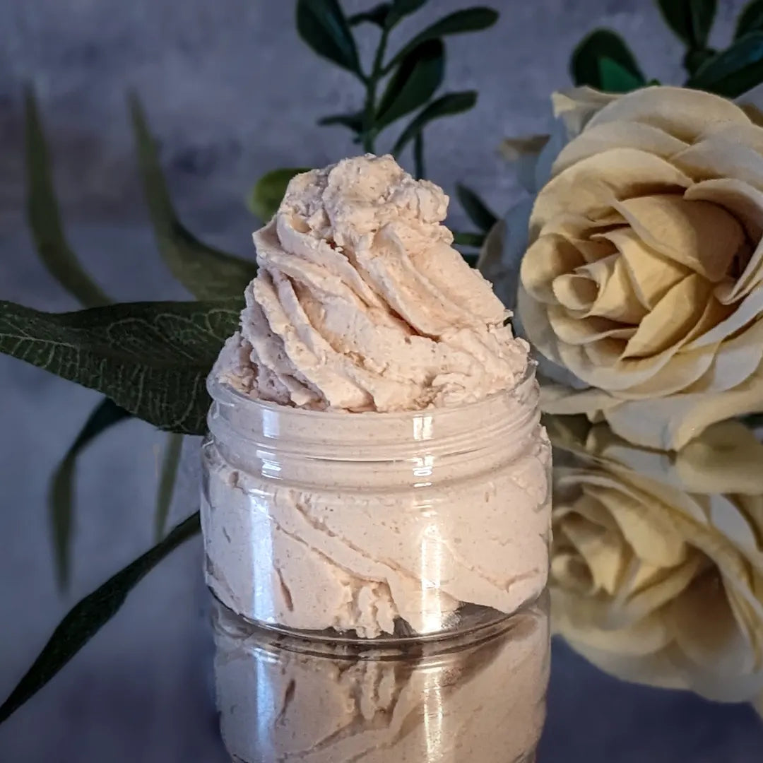 A small jar of light tan whipped body butter, piped like frosting, is on a mirror with a cream colored rose behind the body butter, and in front of a grey concrete background.