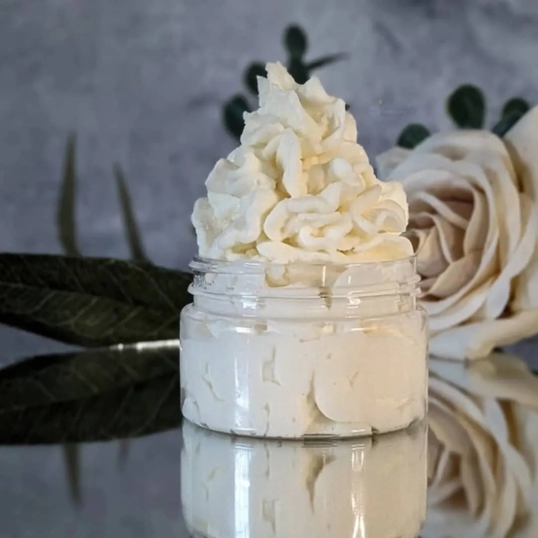cream colored unscented body butter piped like frosting in a clear round jar. The jar is on a mirror, and there's a white rose to the right behind the body butter.