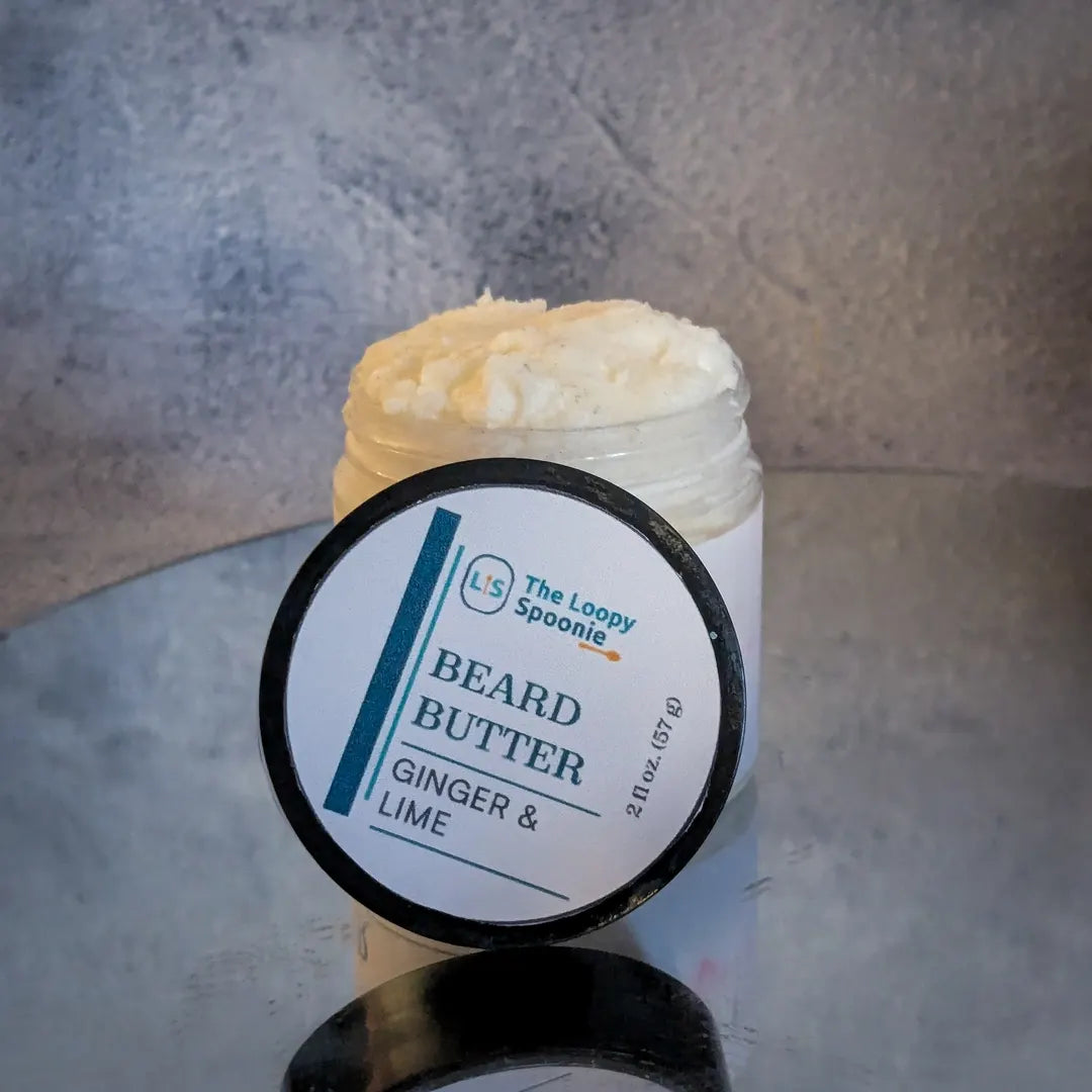 A clear jar of Beard Butter mousse on a mirror, with the lid in front of the jar.
The label on the lid says 'Beard Butter' and the scent 'Ginger & Lime'.
The background is grey concrete/texture