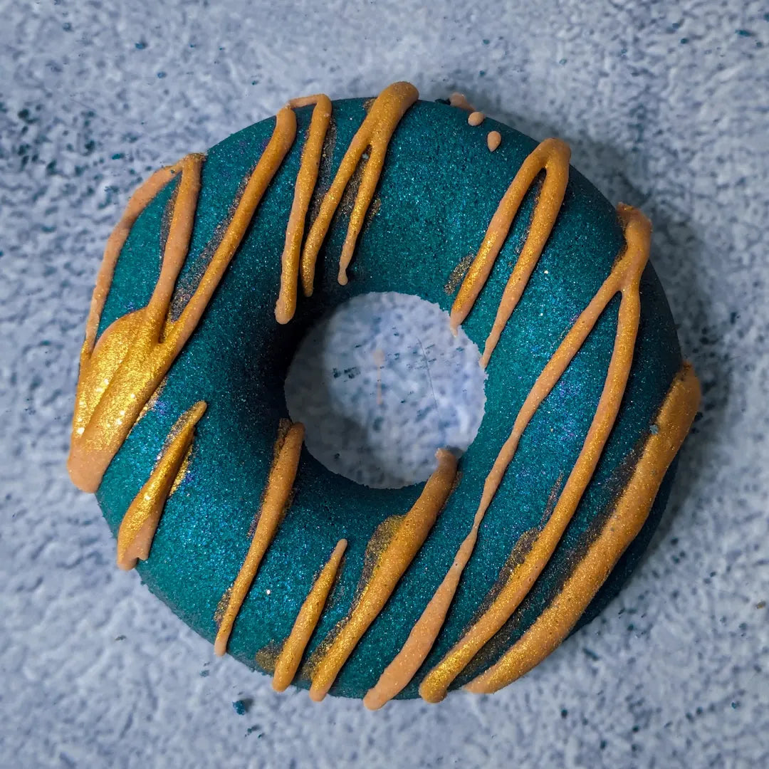 Seafoam green donut bath bomb with gold/yellow cocoa butter drizzled on top.
The bath bomb is on a grey concrete background