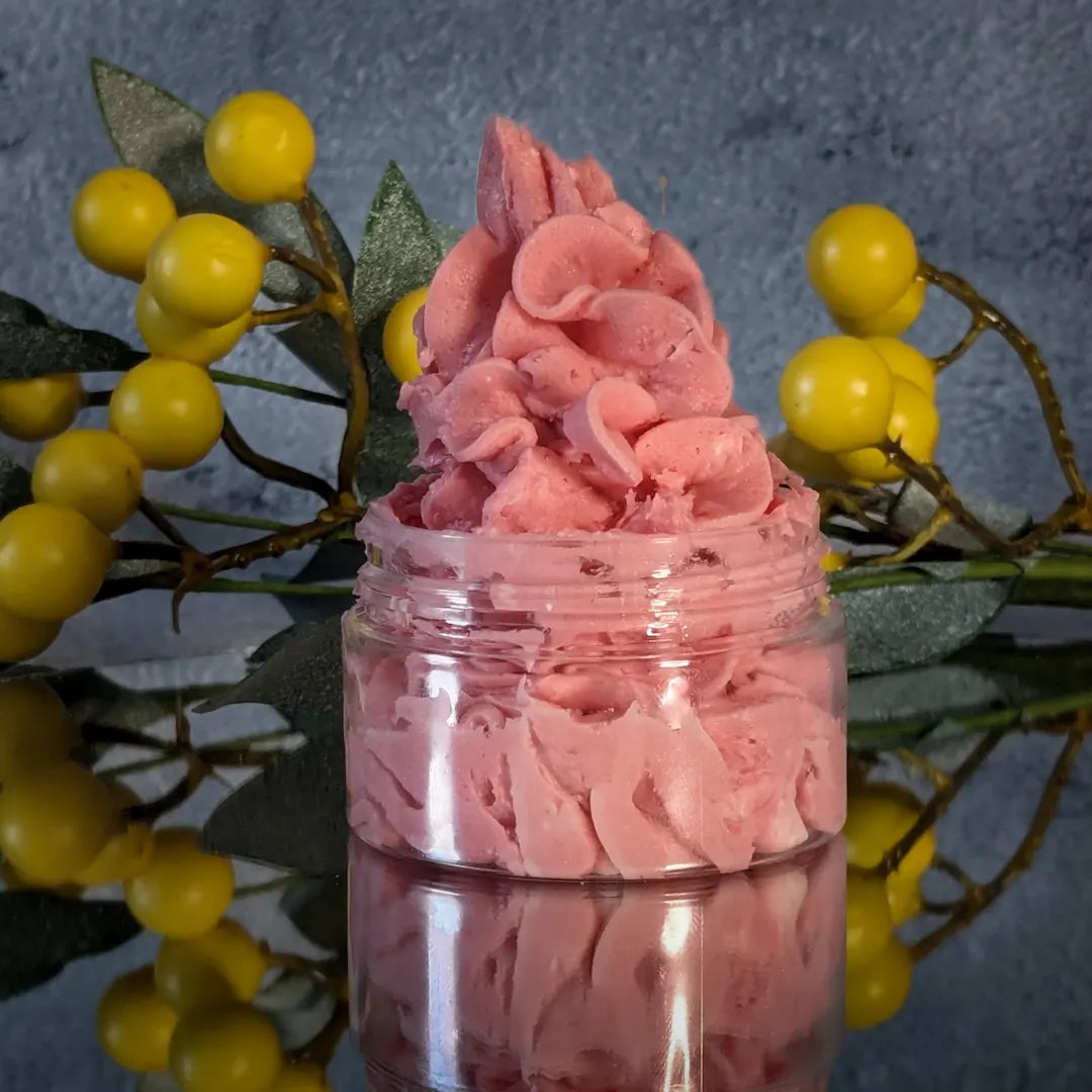 Peachy pink colored whipped body butter piped high like frosting in a small clear jar that sits on a mirror.
There is a sprig of yellow berries & green leaves behind the body butter, in front of a gray concrete background