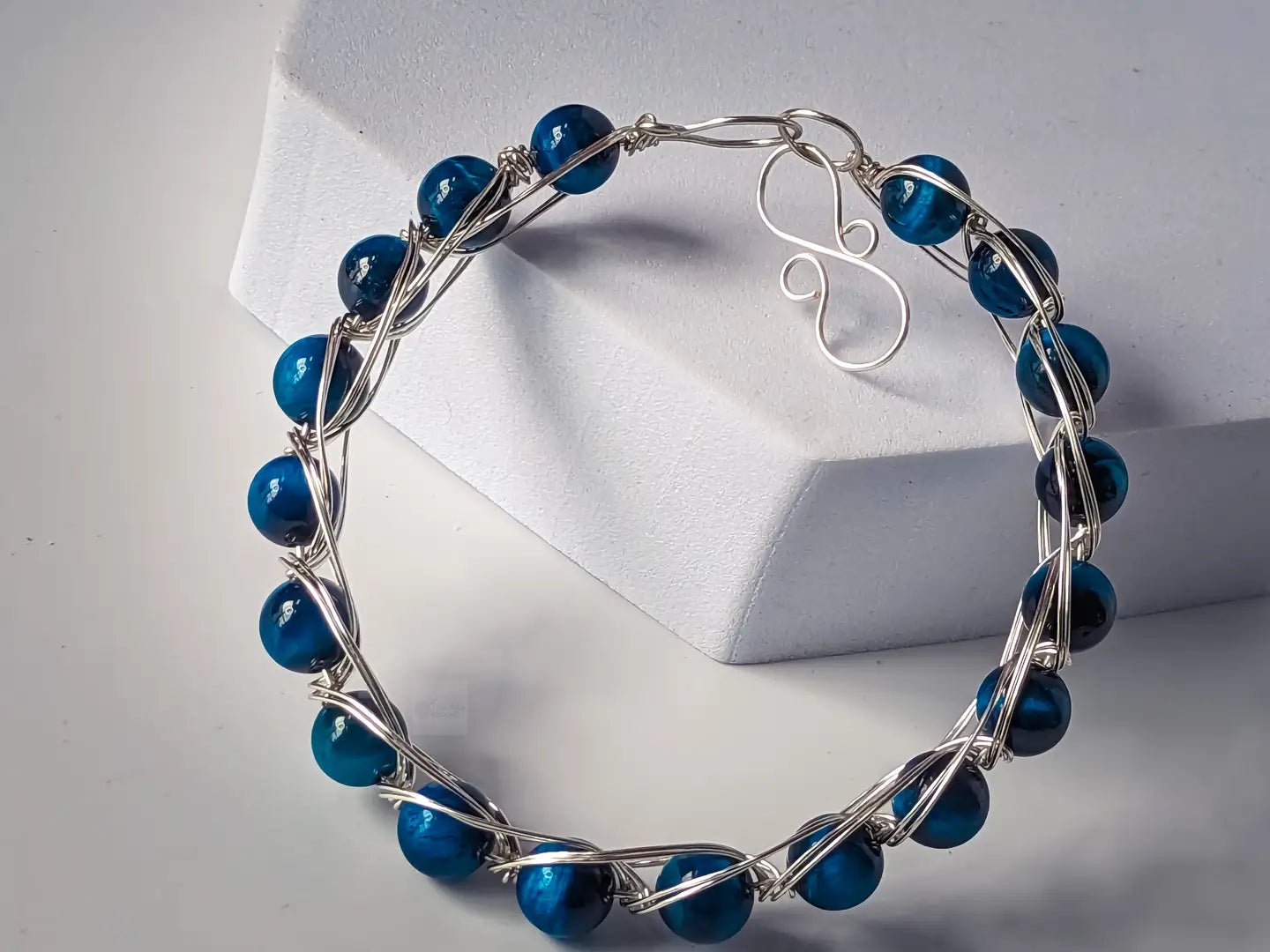 silver wire bracelet with blue tiger eye beads leaning on a white foam block, against a white background.