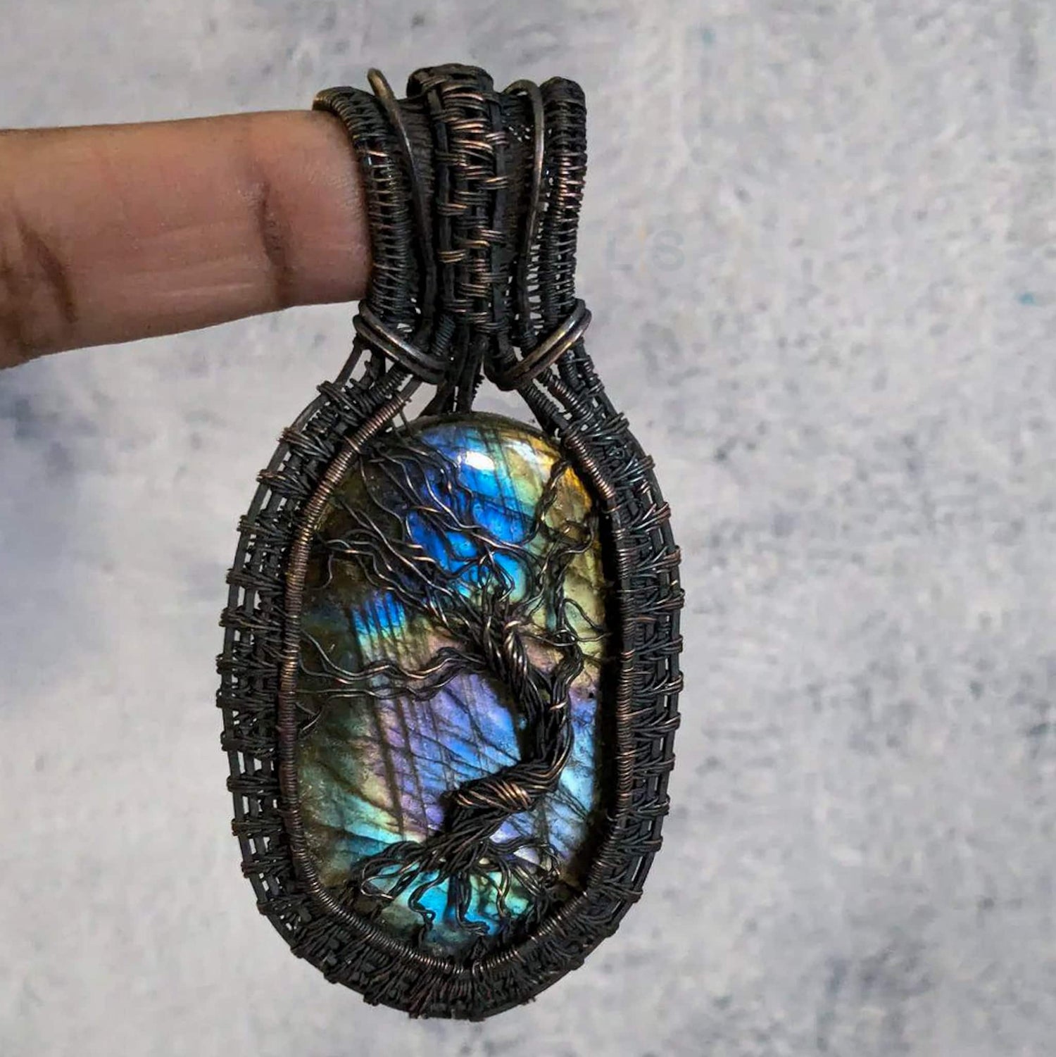 Multiolored large oval labradorite stone with copper wire tree over it and connecting to a copper wire woven frame. The copper wire has been antiqued to be dark brown with brighter copper highlights.