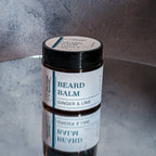 Dark amber jar sitting upright on a mirror against a grey background. The jar has a cream colored wrap around label that says Beard Balm, then below that "Ginger Lime."