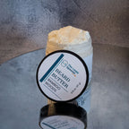 clear jar of whipped beard butter that's upright on a mirror, and has a round lid leaning against the front of the jar. the lid has a round label that has the text: "Beard Butter" then below that "Bamboo Woods".