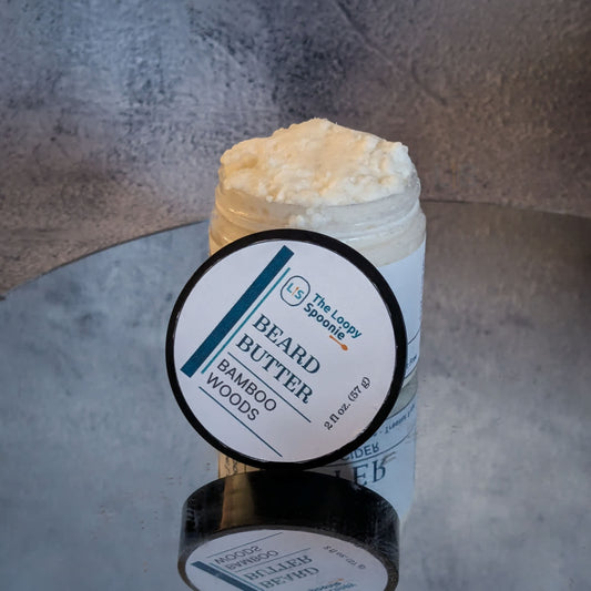 clear jar of whipped beard butter that's upright on a mirror, and has a round lid leaning against the front of the jar. the lid has a round label that has the text: "Beard Butter" then below that "Bamboo Woods".