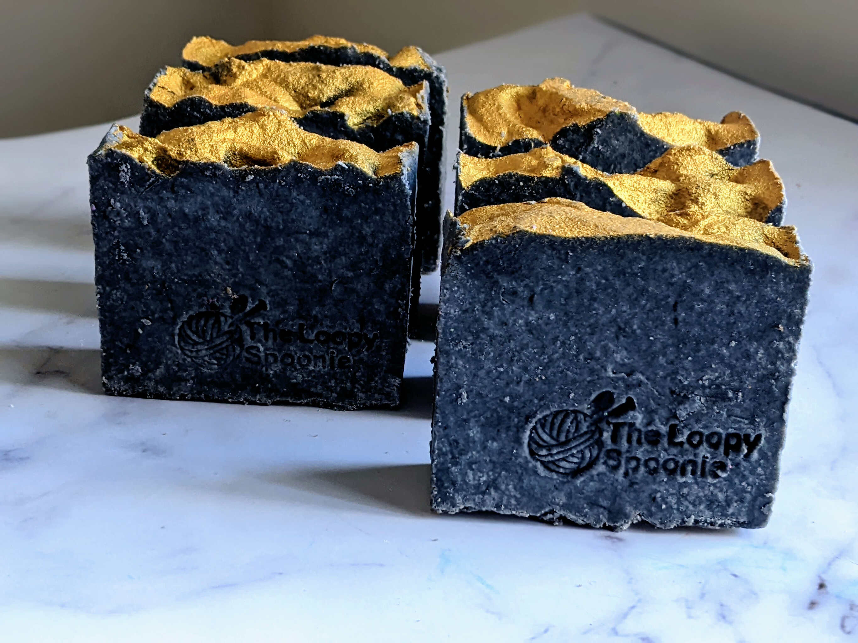 6 rustic black soaps with shiny gold mica soap top and 'The Loopy Spoonie' stamped on the front of the soaps. The Soaps are upright and lined up in 2 rows of 3 on white marble surface.