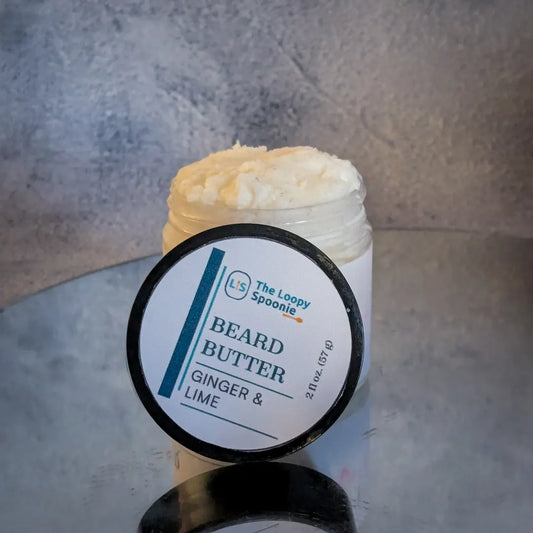 A clear jar of Beard Butter  mousse on a mirror, with the lid in front of the jar. 

The label on the lid says 'Beard Butter' and the scent 'Ginger & Lime'.

The background is grey concrete/texture