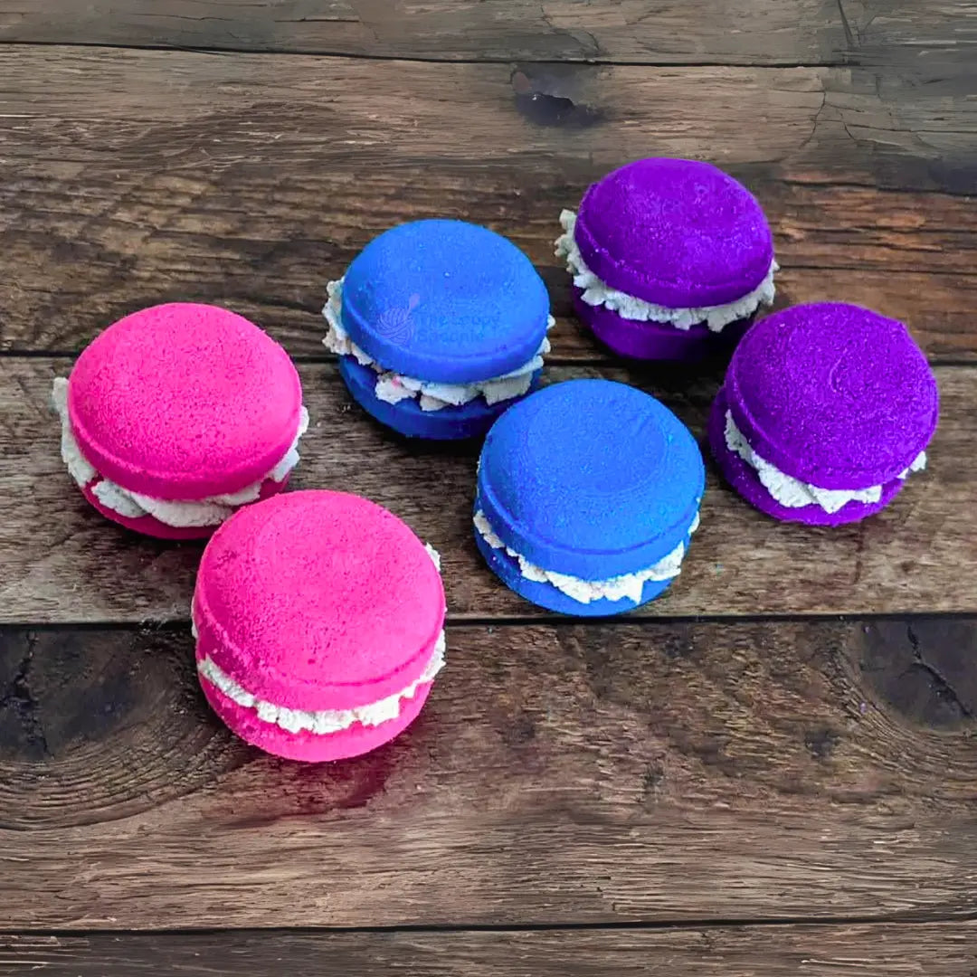 6 macaron-shaped bath bombs in pink, blue, and purple colors with a white bubble frosting design on a wooden surface.