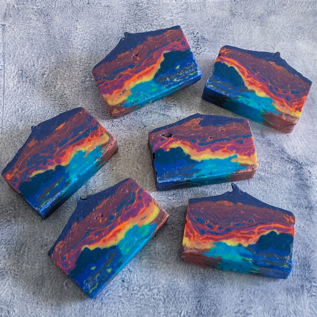 6 rectangular landscape soap bars designed to resemble a marbled neon ocean sunset. The soaps are haphazardly arranged on a grey textured background.