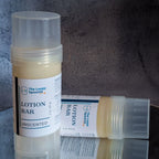 Two lotion bar sticks on a mirror against grey concrete backdrop. 

One lotion bar is upright, the second is behind and laying sideways