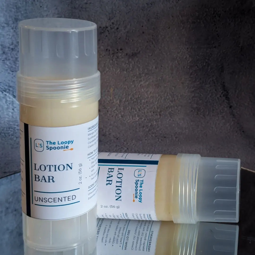 Two lotion bar sticks on a mirror against grey concrete backdrop. 

One lotion bar is upright, the second is behind and laying sideways