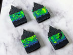 4 frosted soap bars meant to resemble the northern lights with neon green & blue swirls above a black cityscape at the bottom of the soap. The soap bars laying flat on a marble surface