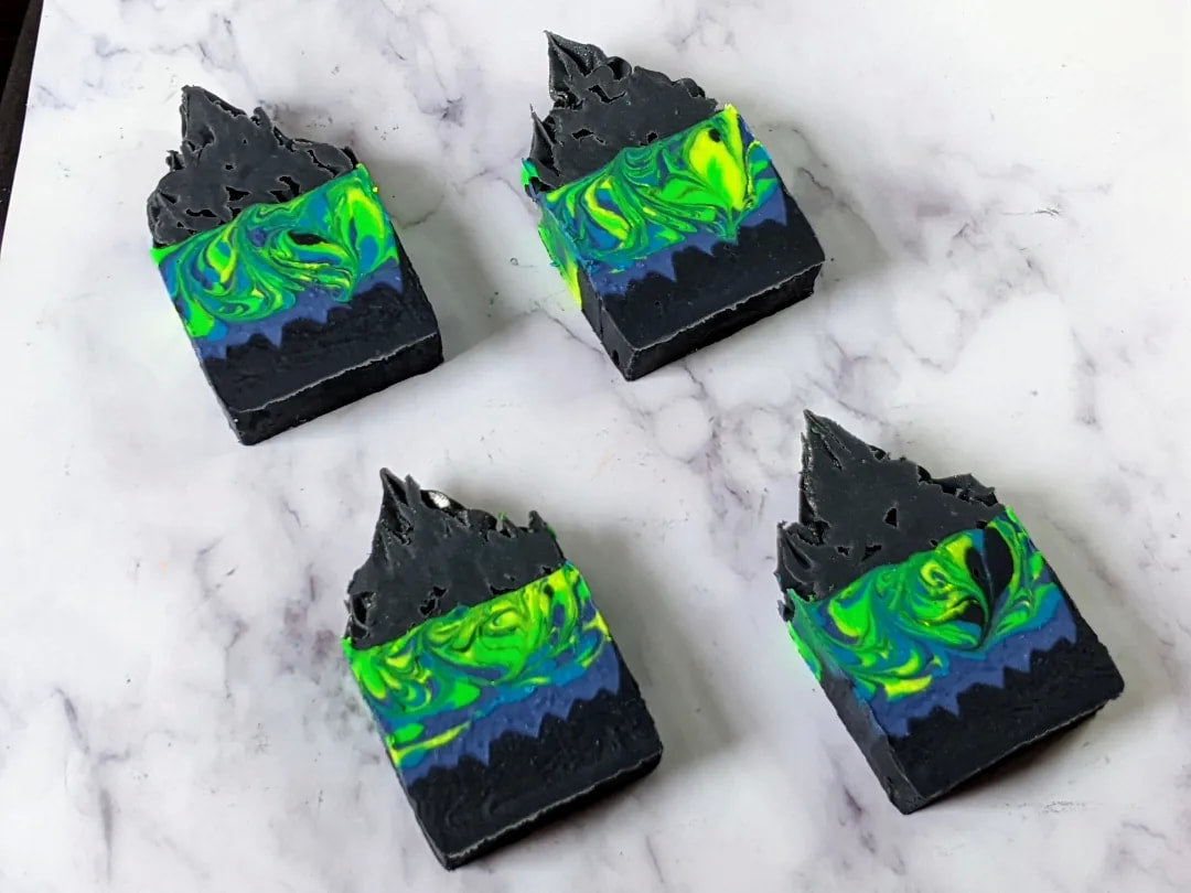 4 frosted soap bars meant to resemble the northern lights with neon green & blue swirls above a black cityscape at the bottom of the soap. The soap bars laying flat on a marble surface