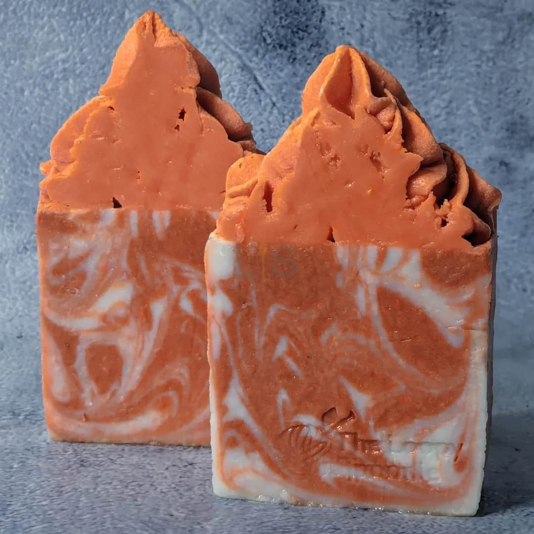 Two bars of orange & cream colored drop swirl soap in a marbled-like pattern, and orange soap frosting piped on top. Both soaps are upright on a gray concreate background