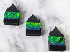 3 black, neon green & blue frosted soap bars laying flat in a diagonal line on a marble surface