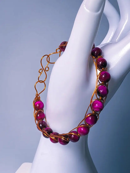 A wire wrapped bracelet featuring pink tiger eye beads is displayed on the thumb of a white mannequin hand in front of a light grey/blue background.