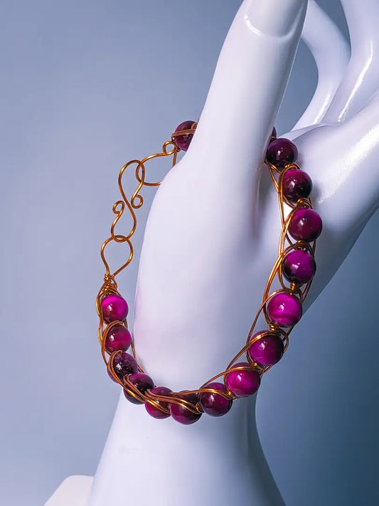 A wire wrapped bracelet featuring pink tiger eye beads is displayed on the thumb of a white mannequin hand in front of a light grey/blue background.