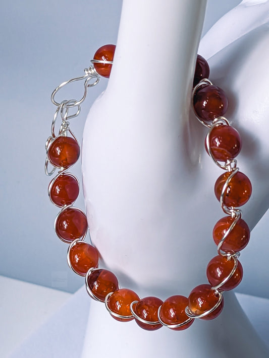Red & orange dragon vein agate beads wrapped/accented by silver plated copper to form a bracelet. The bracelet is hanging on the thumb of a white mannequin hand in front of a grey backdrop.