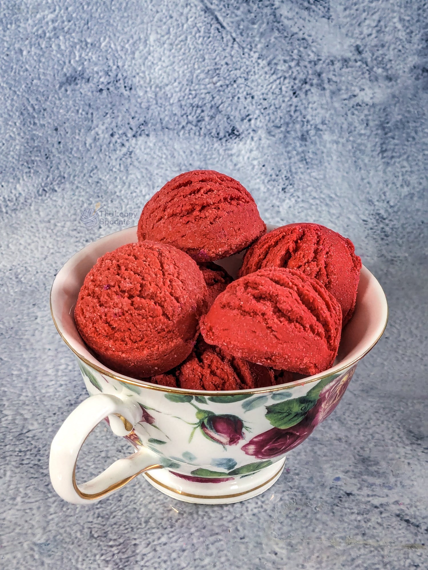 Several red strawberry scented mini bubble bath scoops in a floral teacup against a grey textured background.