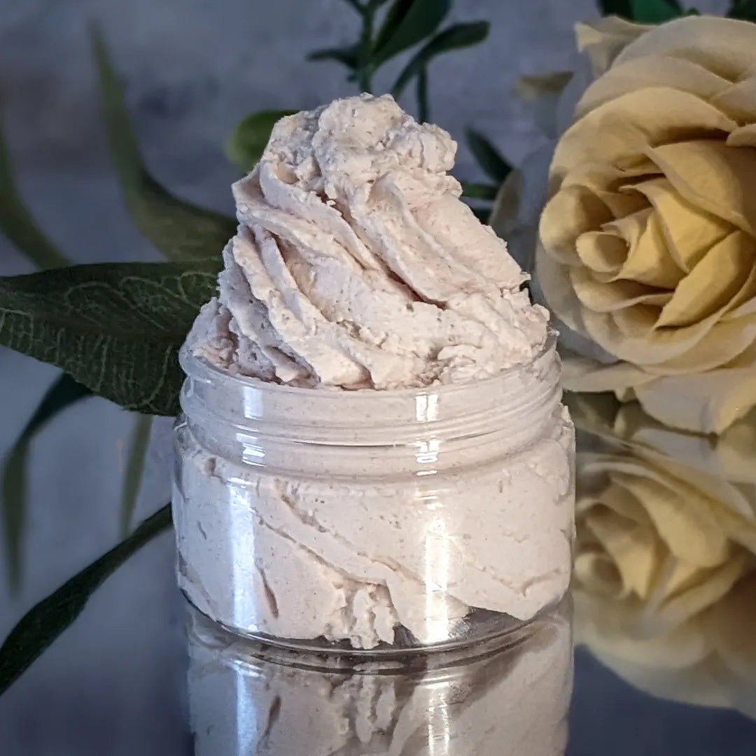 A small jar of cream colored body butter, that's been piped like frosting. 
The jar is on a mirror with a yellow rose to the right behind the body butter, and in against a grey concrete background.