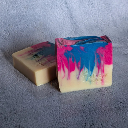 2 cream bars of soap with bright pink & blue stripes that look like dripping paint from the top of the soap bar.

One soap is lying flat, while the other soap is upright in front of the 1st soap. Both soaps are on a grey concrete background