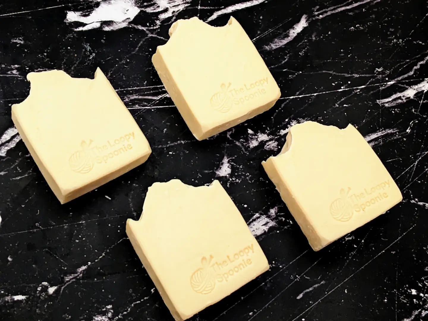 4 cream colored bastille-castile soaps on a black marble background. Each bar has 'The Loopy Spoonie' embossed/stamped in the bottom right of the soap bar