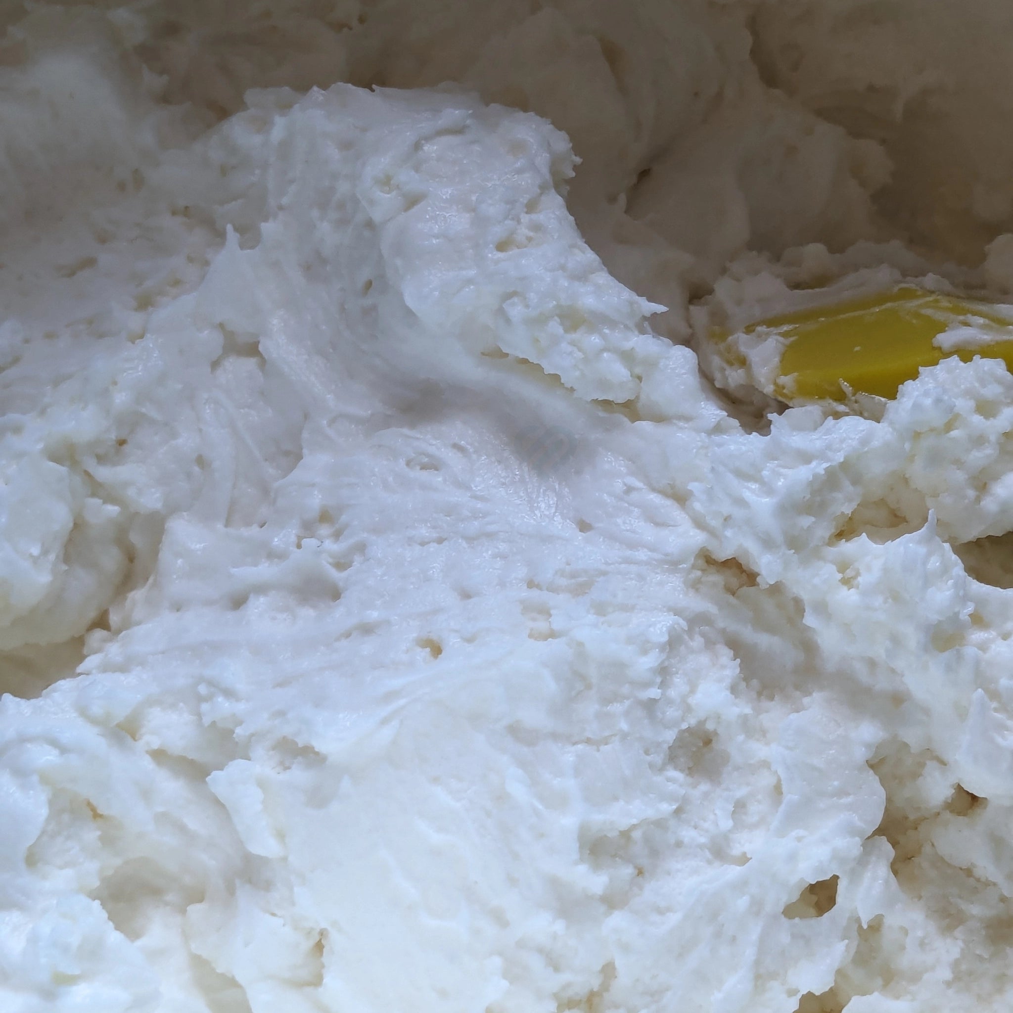 A close-up of thick, fluffy white whipped body butter butter with a yellow silicone spatula visible on the right side.